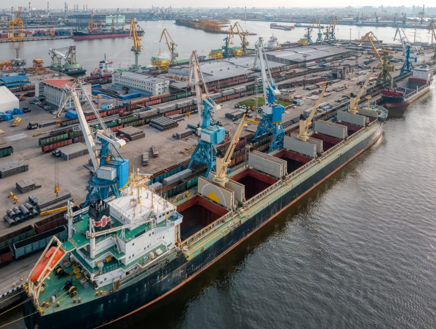 A photo of port with boats, cranes and buildings. Port cranes load various cargoes into the hold of the ship. Goods delivery by sea. On the background railway wagons. Global economic.